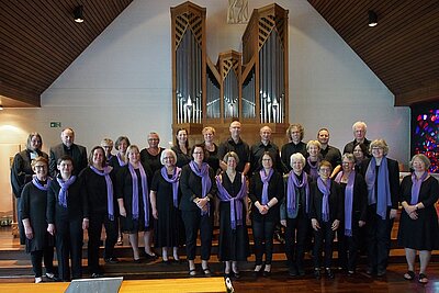 Der versammelte Chor in der Erlöserkirche. Der Chor, ganz in schwarz gekleidet, die Damen mit lila Schal akzentuiert, steht dicht zusammen am Altar. Im Hintergrund ist die Orgel zusehen. 