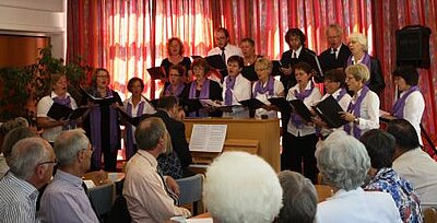 Der Chor bei einer Aufführung im Gemeindehaus. Im Vordergrund zu sehen ist das Publikum und Thomas Schulze-Athens am Klavier. Auf der Bühne im Hintergrund ist der Chor zu sehen.